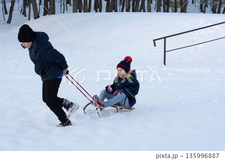 A child pulls a sled carrying another child through snowy park landscape. A child pulls a sled carrying another child through snowy park landscape. 135996887
