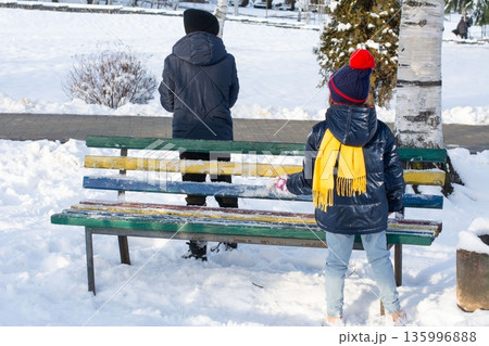 Two children take a break from playing in the snow, sitting on opposite ends of a colorful park bench. 135996888