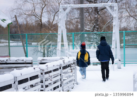 Two figures stroll through a snow-covered park, passing a swing set and snow-laden planters. Two figures stroll through a snow-covered park, passing a swing set and snow-laden planters. 135996894