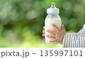 Mother or caregiver feeding infant with bottle in natural daylight at outdoor setting 135997101