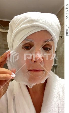 A woman in the bathroom adjusts a cloth care mask on her face.  135997109