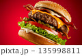 Big burger with bacon, cheese, lettuce, tomato, and sauce on sesame seed bun against red background 135998811