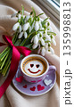Fresh coffee with heart design next to a bouquet of flowers on a soft cloth near a window 135998813
