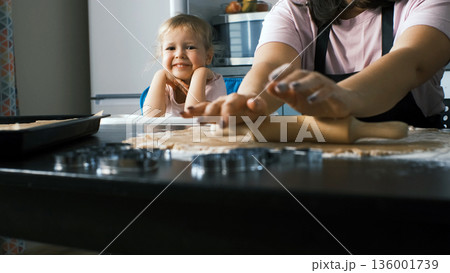 Smiling young girl watches as ginger cookie dough is rolled on the kitchen table, observing her mother during a calm and peaceful family baking moment 136001739