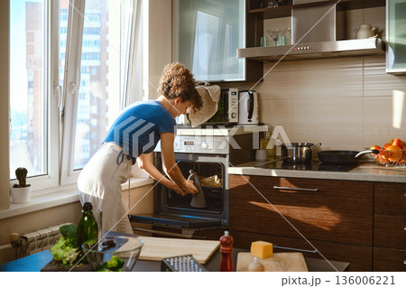 Woman is taking out a tray from the oven in a kitchen Woman is taking out a tray from the oven in a kitchen 136006221