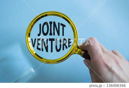 Joint Venture symbol. Magnifying Glass with words Joint Venture. Businessman hand. Beautiful blue background. Business and Joint Venture concept. Copy space. 136006366
