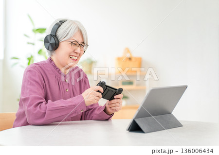 家のリビングでヘッドホンをしてゲームをするグレイヘアのアジア人女性 136006434