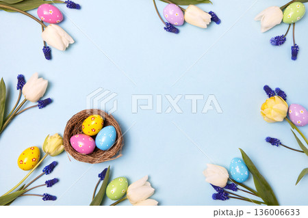Easter colored painted eggs in bird nest with yellow daffodils, white tulips, and blue muscari on blue background with space for text and greetings. Easter and spring concept. Spring background Easter colored painted eggs in bird nest with yellow daffodils, white tulips, and blue muscari on blue background with space for text and greetings. Easter and spring concept. Spring background 136006633