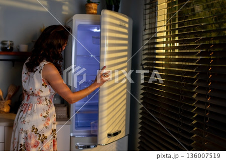 Evening kitchen activity, Lady opens fridge late at night, Woman in floral outfit seeks jar in darkness, Night example of woman retrieving item from cold storage 136007519