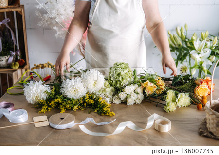 Florist arranges fresh blossoms on paper covered table in bright studio. Artisan craftsmanship, small business growth, floral design. 136007735