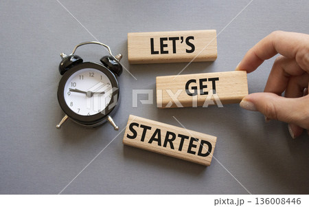 Lets get started symbol. Concept words Lets get started on wooden blocks. Businessman hand. Beautiful grey background with alarm clock. Business and Lets get started concept. Copy space. Lets get started symbol. Concept words Lets get started on wooden blocks. Businessman hand. Beautiful grey background with alarm clock. Business and Lets get started concept. Copy space. 136008446