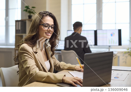 Happy businesswoman using laptop and pencil at bright office workstation and graphs 136009046