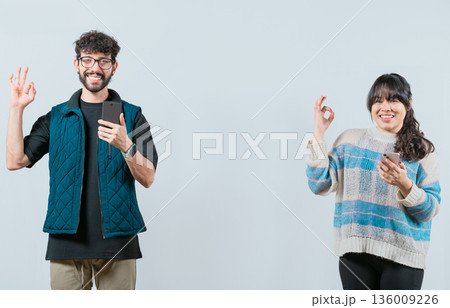 Smiling couple holding a cell phone and gesturing OK, isolated. Happy young couple holding phone gesturing OK 136009226