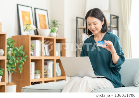 リビングでクレジットカートとパソコンを見るミドルの女性 リビングでクレジットカートとパソコンを見るミドルの女性 136011706