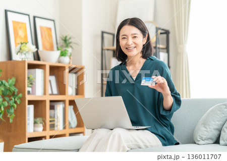 リビングでクレジットカートとパソコンを見るミドルの女性 リビングでクレジットカートとパソコンを見るミドルの女性 136011707