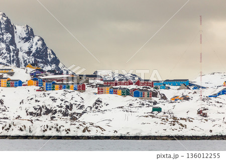 Wide coastal panorama of Sisimiut with colorful Inuit houses, snow covered terrain and steep arctic mountains rising above the calm fjord, Greenland 136012255