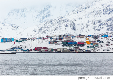 Wide coastal panorama of Sisimiut with colorful Inuit houses, snow covered terrain and steep arctic mountains rising above the calm fjord, Greenland 136012256