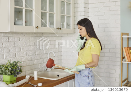Woman standing in kitchen with plunger talking by mobile phone with plumber trying to unclog a sink 136013679