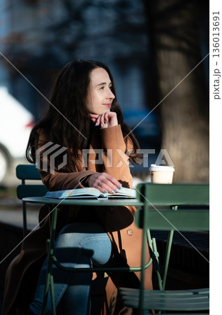 Contemplative afternoon at a green cafe table, a woman enjoys coffee while lost in thought in a charming urban street setting, capturing the essence of quiet moments 136013691
