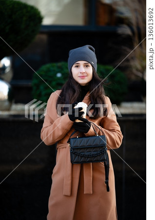 Charming young woman enjoys a warm beverage while standing against a well-manicured backdrop on a crisp autumn day in a bustling city 136013692