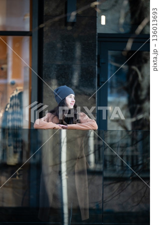 Young woman enjoying the urban view from a modern glass balcony on a chilly day, wrapped in cozy attire and a warm beanie while reflecting on her surroundings 136013693