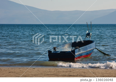 Silhouetted fishing boat on the shore of Vlora, Albania, captured on August 24, 2025. The backlit scene shows shimmering sunlight on the sea, gentle waves, and a swimmer in the background, creating a  136016048