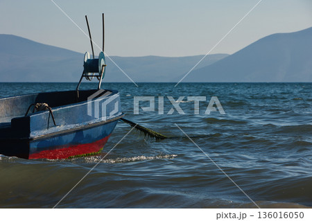 Silhouetted fishing boat on the shore of Vlora, Albania, captured on August 24, 2025. The backlit scene shows shimmering sunlight on the sea, gentle waves, and a swimmer in the background, creating a  136016050