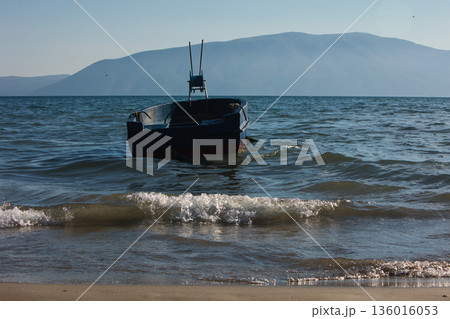 Silhouetted fishing boat on the shore of Vlora, Albania, captured on August 24, 2025. The backlit scene shows shimmering sunlight on the sea, gentle waves, and a swimmer in the background, creating a  136016053