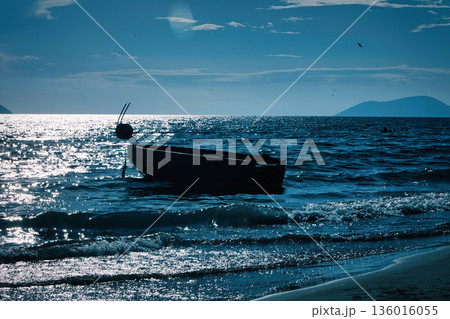Silhouetted fishing boat on the shore of Vlora, Albania, captured on August 24, 2025. The backlit scene shows shimmering sunlight on the sea, gentle waves, and a swimmer in the background, creating a  136016055