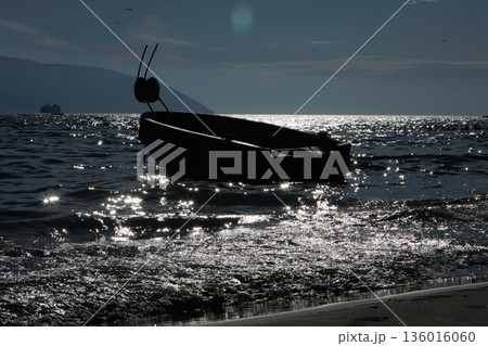 Silhouetted fishing boat on the shore of Vlora, Albania, captured on August 24, 2025. The backlit scene shows shimmering sunlight on the sea, gentle waves, and a swimmer in the background, creating a  136016060