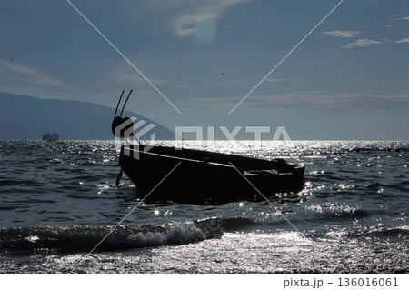 Silhouetted fishing boat on the shore of Vlora, Albania, captured on August 24, 2025. The backlit scene shows shimmering sunlight on the sea, gentle waves, and a swimmer in the background, creating a  136016061