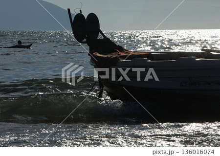 Silhouetted fishing boat on the shore of Vlora, Albania, captured on August 24, 2025. The backlit scene shows shimmering sunlight on the sea, gentle waves, and a swimmer in the background, creating a  136016074