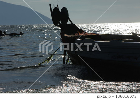 Silhouetted fishing boat on the shore of Vlora, Albania, captured on August 24, 2025. The backlit scene shows shimmering sunlight on the sea, gentle waves, and a swimmer in the background, creating a  136016075