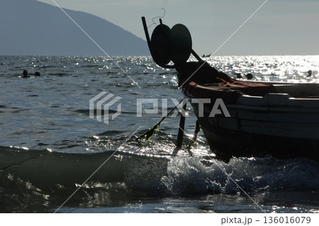 Silhouetted fishing boat on the shore of Vlora, Albania, captured on August 24, 2025. The backlit scene shows shimmering sunlight on the sea, gentle waves, and a swimmer in the background, creating a Silhouetted fishing boat on the shore of Vlora, Albania, captured on August 24, 2025. The backlit scene shows shimmering sunlight on the sea, gentle waves, and a swimmer in the background, creating a 136016079