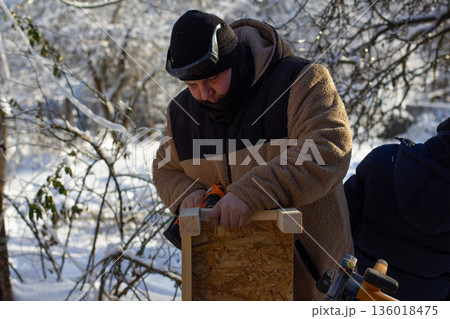 Man in warm winter clothing engaging in a diy project, constructing a wooden birdhouse outdoors during a cold, snowy winter day, symbolizing hobby, craftsmanship, and caring for nature. 136018475