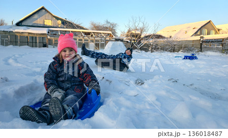 Young children laughing and enjoying themselves while sledding down a snowy slope, experiencing the joy of outdoor winter play in a residential backyard with houses in the background. Young children laughing and enjoying themselves while sledding down a snowy slope, experiencing the joy of outdoor winter play in a residential backyard with houses in the background. 136018487