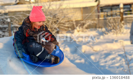A child rides down a hill in a sleigh. Child enjoying winter holiday outdoors, riding down snowy slope on blue plastic sled with joyful expression on face under golden sunlight. 136018488