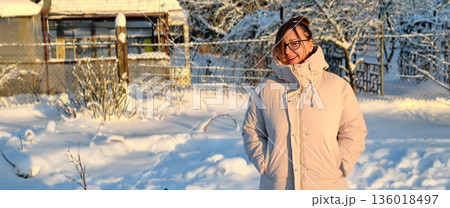A woman stands in a snowy courtyard wearing a warm down jacket and sunglasses, smiling and looking away. On a bright, cold winter day, the woman enjoys the peaceful scenery outside. 136018497