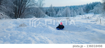 Child wearing a pink hat having fun sledding down a snowy slope, surrounded by a serene winter landscape with frost-covered trees creating a peaceful and playful childhood scene. 136018498