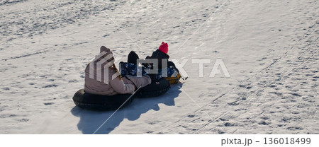 A family enjoys a sunny winter day tubing down a snowy hill. Children and parents laugh together as they glide down the slopes, bonding, and having fun outdoors in the frosty landscape. 136018499