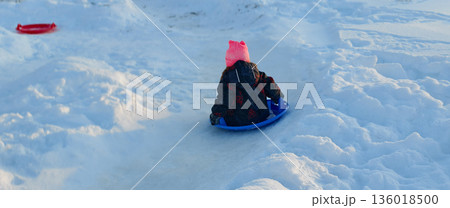 A girl in a pink hat and winter coat rides down a snowy slope on a blue sled, shot from behind. The child laughs and races down the icy hill against the backdrop of a bright, crisp winter landscape. A girl in a pink hat and winter coat rides down a snowy slope on a blue sled, shot from behind. The child laughs and races down the icy hill against the backdrop of a bright, crisp winter landscape. 136018500