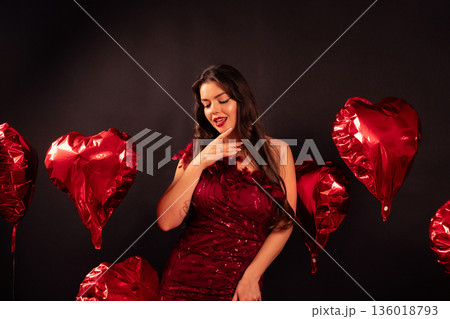 Valentines day celebration with woman wearing red dress surrounded by heart balloons in studio 136018793