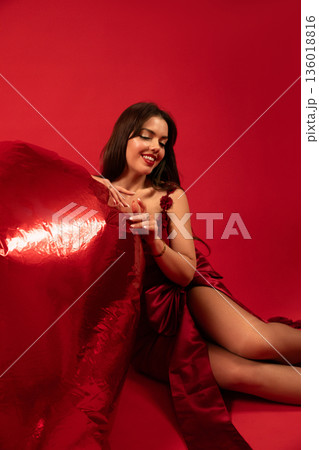 Beautiful woman sitting on red background holding a large heart-shaped decoration for Valentines Day celebration Beautiful woman sitting on red background holding a large heart-shaped decoration for Valentines Day celebration 136018816