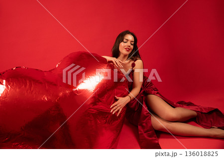 Woman sits with large red heart while wearing a red dress in a studio setting for Valentines Day celebration Woman sits with large red heart while wearing a red dress in a studio setting for Valentines Day celebration 136018825