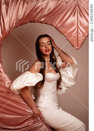 Beautiful woman in a white dress posing with pink fabric on Valentines Day setting in a studio Beautiful woman in a white dress posing with pink fabric on Valentines Day setting in a studio 136018849