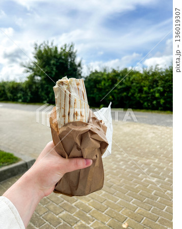 Woman holds shawarma in hand on street. Sheet food. Eating Kebab with chicken Woman holds shawarma in hand on street. Sheet food. Eating Kebab with chicken 136019307