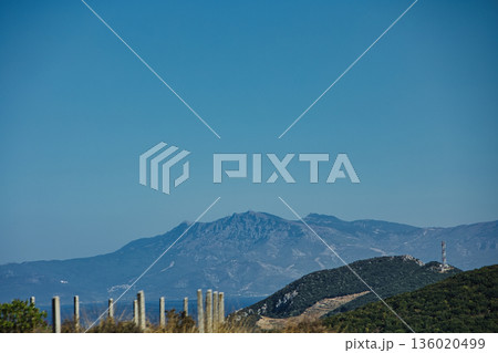 Mountain range rising above a calm blue sea under a clear sky, forming a minimalistic coastal landscape. Serene natural scenery captured on August  2025. 136020499