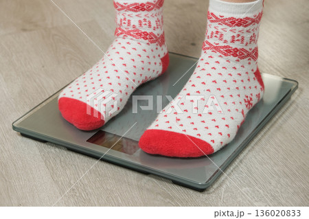 Feet in Cozy Christmas Socks Standing on Digital Bathroom Scales, New Year's Resolution Concept. Feet in Cozy Christmas Socks Standing on Digital Bathroom Scales, New Year's Resolution Concept. 136020833