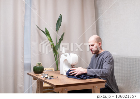 A bald man of 40 years old in a gray T-shirt sits at a table and sews on a sewing machine at home 136021557