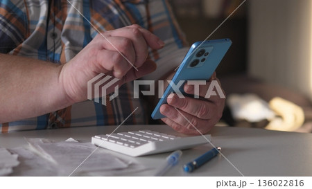 A man sits at a table using a smartphone with one hand while the other hand rests on a calculator. Papers are spread out on the table in a bright room during the afternoon. A man sits at a table using a smartphone with one hand while the other hand rests on a calculator. Papers are spread out on the table in a bright room during the afternoon. 136022816